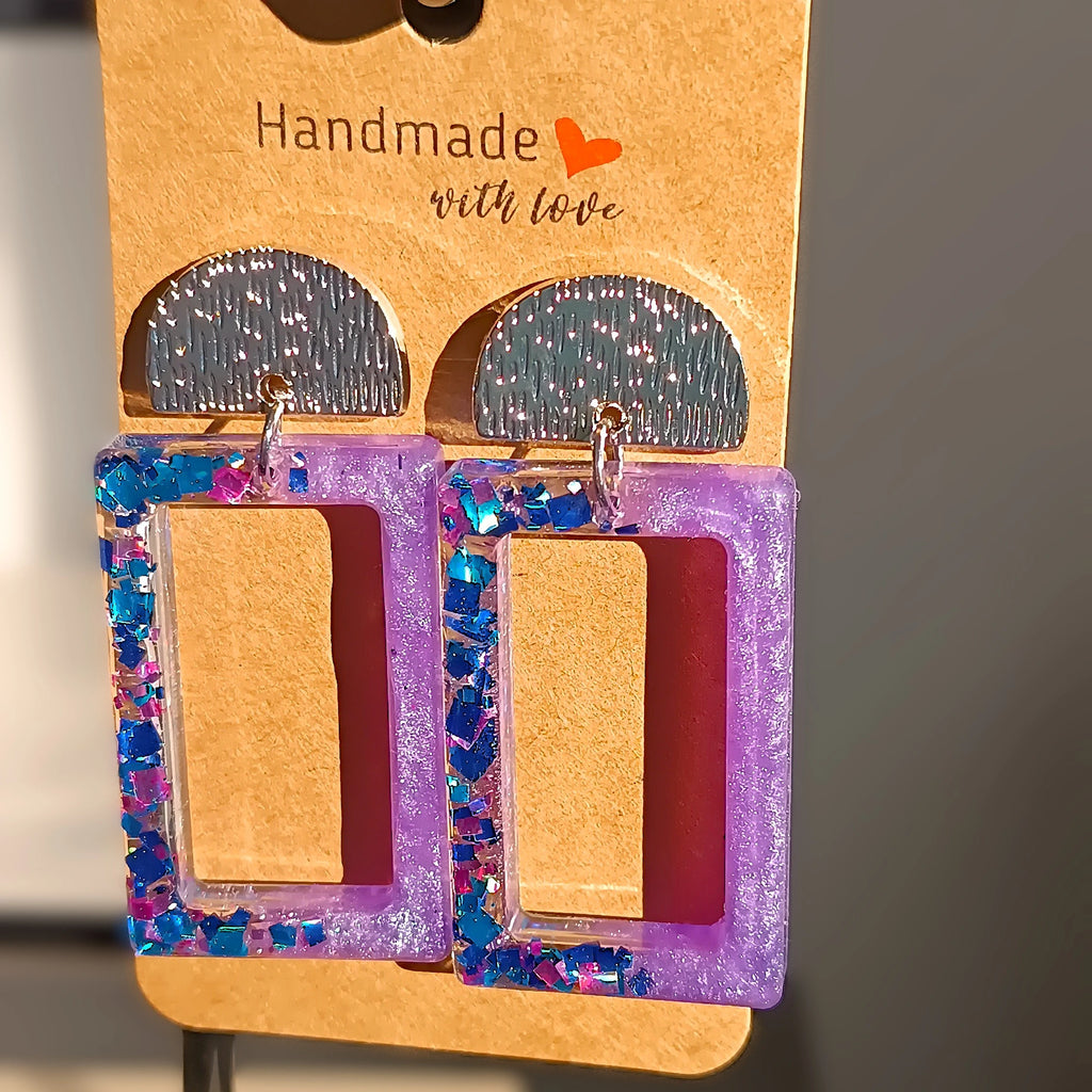 Boucles d’oreilles rectangulaires en résine UV  mauve avec paillettes bleues et mauves, bijoux faits main, posées sur un carton de présentation