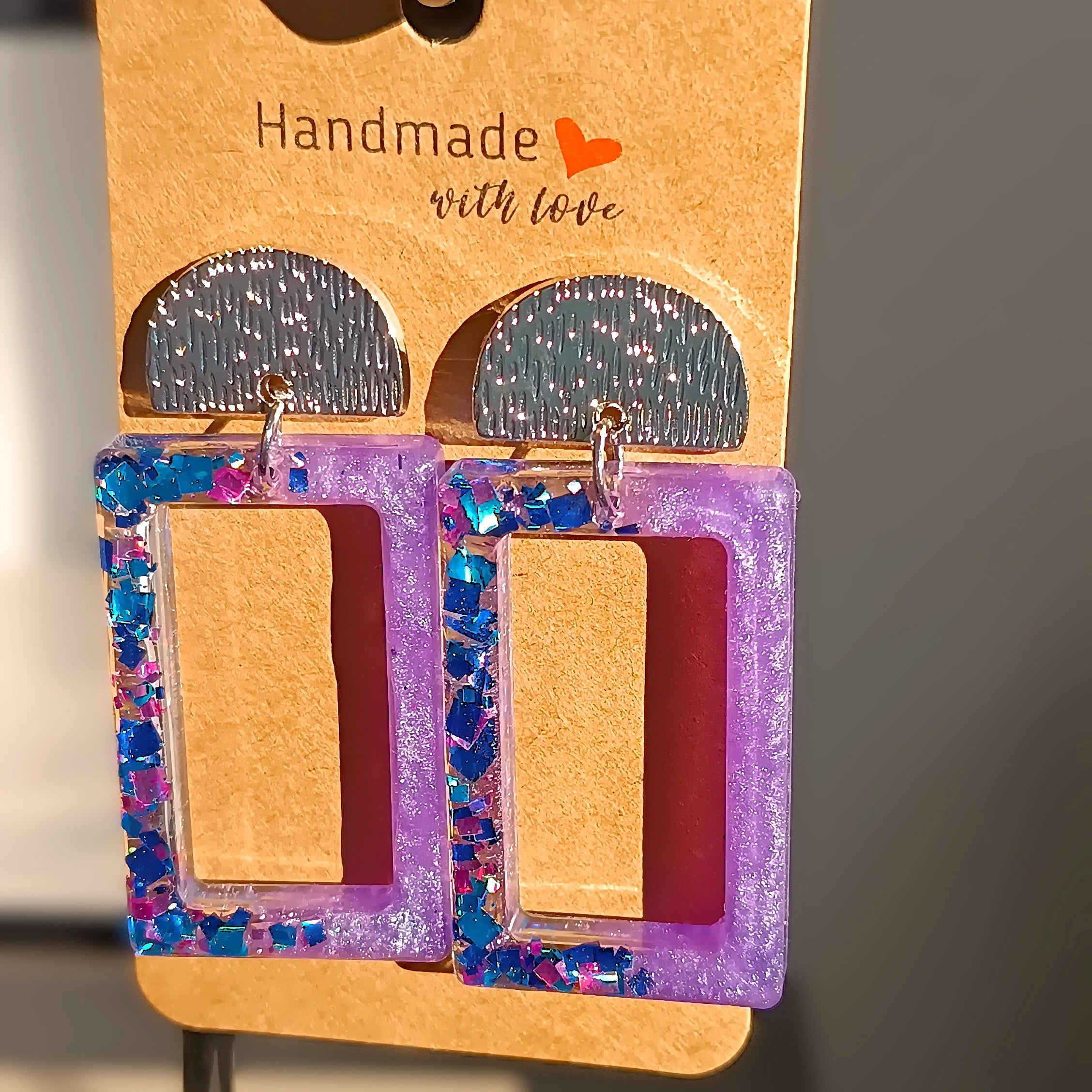 Boucles d’oreilles rectangulaires en résine UV  mauve avec paillettes bleues et mauves, bijoux faits main, posées sur un carton de présentation