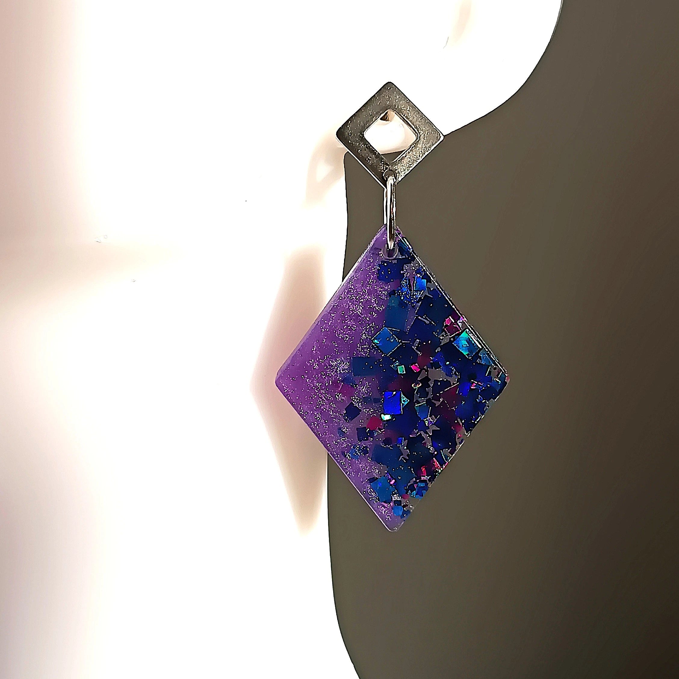 Boucles d’oreilles losange en résine mauve avec des paillettes bleues et mauves, portées sur un mannequin.